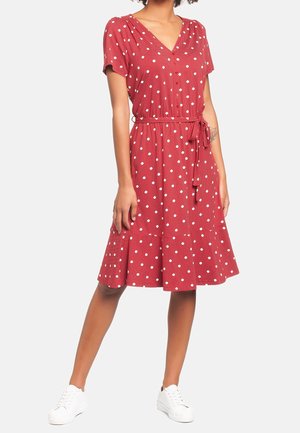 Frau trägt ein rotes, knielanges Kleid mit weißen Punkten, kurzen Ärmeln, V-Ausschnitt, gebundener Taille und weißen Sneakers, steht mit einem Bein nach vorne.