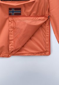 Oranje jack met een gaaszak, voorzien van een rechthoekig patch met een vlagontwerp. Glad materiaal met een ritssluiting.