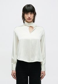 Kremowa satynowa bluzka z wysokim kołnierzem, z przodu posiada wycięcie w kształcie łezki, długie rękawy i lekko zaokrąglony dół. W połączeniu z czarnymi spodniami.