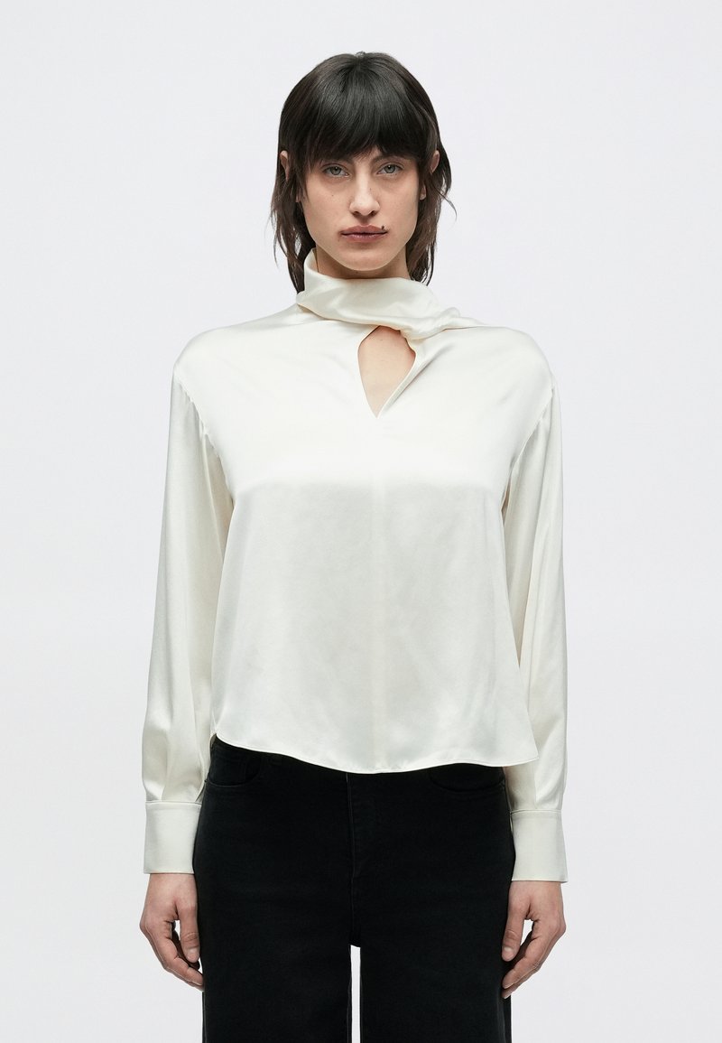 Kremowa satynowa bluzka z wysokim kołnierzem, z przodu posiada wycięcie w kształcie łezki, długie rękawy i lekko zaokrąglony dół. W połączeniu z czarnymi spodniami.