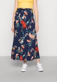 Blomstrande lång kjol i marinblått tyg, med mönster av röda och gröna blommor, elastisk midja med snöre, i kombination med vita sneakers.