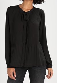 Blouse noire plissée avec un col rond, fermeture à nouer au niveau du col et manches longues. Tissu léger avec une texture douce.