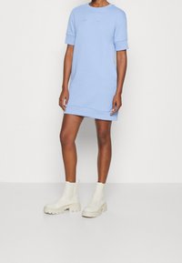 Ljusblå bomullsklänning med korta ärmar, prydd med broderad logotyp. Matchas med vita ankelstövlar för en avslappnad look. Minimalistisk design.