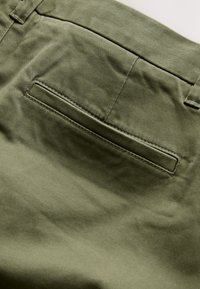 Pantalon vert olive en gros plan montrant une poche arrière passepoilée avec coutures visibles et détail passants de ceinture.