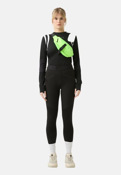 Black long-sleeve top, black high-waisted pants with zippers, green crossbody bag, white sneakers, and white socks.