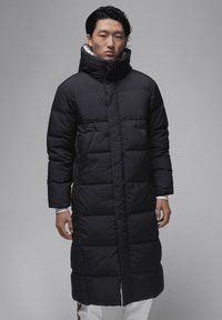 Lange sort pufferjakke med høj krave, der har et kviltet design og en let skinnende overflade, båret over hvide tøj.