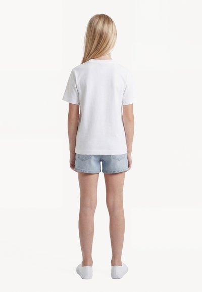 Παιδί με μακριά ξανθά μαλλιά, γυρισμένο προς τα πίσω, φοράει ένα απλό λευκό μπλουζάκι, ανοιχτόχρωμα τζιν σορτς και λευκά παπούτσια χωρίς κορδόνια.