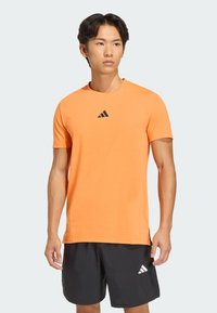 Camiseta deportiva naranja de manga corta con escote redondo y logo negro de Adidas en el pecho, confeccionada con una tela ligera y suave.