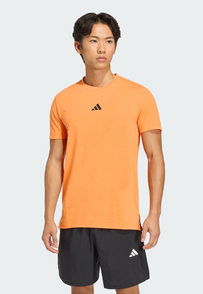 Camiseta deportiva naranja de manga corta con escote redondo y logo negro de Adidas en el pecho, confeccionada con una tela ligera y suave.