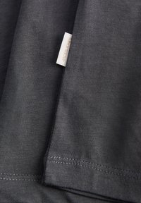 Σκούρο γκρι βαμβακερό ύφασμα με μια μικρή άσπρη ραμμένη ετικέτα που δείχνει αχνό κείμενο, κοντινή λήψη της διπλωμένης άκρης ένδυσης και ραφής.