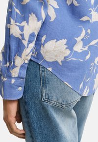 Camicia azzurro chiaro con motivo floreale bianco, con polsini a bottone e spacco laterale, abbinata a jeans in denim chiaro. Tessuto testurizzato, design casual.