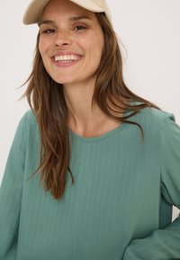 Maglia a maniche lunghe color teal con strisce verticali, scollatura rotonda e texture morbida; abbinata a un cappello beige chiaro.