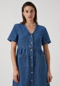 Denimkjole med korte ermer, V-hals, knappestolpe og to frontlommer. Blått stoff med rynket midje og sømdetaljer.