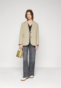 Blazer oversize beige con bottoni marroni, abbinato a un top nero e jeans a palazzo grigi. Accessorio: piccola borsa a tracolla con motivo verde.