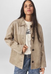 Beige suède jacket met een gezwikte kraag, knoopsluitingen en zijzakken. Draag het over een licht t-shirt en combineer het met versleten denim.