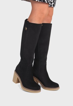 Bottes en daim noir montant jusqu'au genou avec des talons beiges épais, portées par une personne debout, avec une jupe à motifs visible au-dessus des bottes.