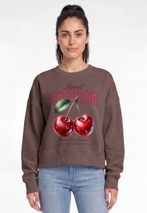 Jonge vrouw die een bruine trui draagt met een grote afbeelding van twee kersen en de tekst "Sweet Temptations", staand voor een witte achtergrond.
