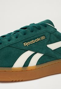 Zelená semišová teniska s bílými doplňky, zdobená zlatým logem Reebok, s texturovanou podrážkou z gumy v přírodní barvě a klasickým šněrováním.