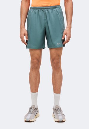 Mężczyzna ubrany w turkusowe sportowe szorty Nike, białe skarpety za kostkę i szare buty do biegania Nike, stoi na jednolitym tle.