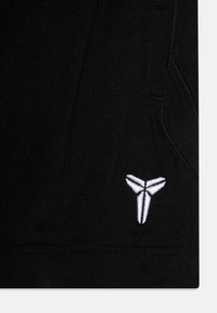 Schwarzer Sportstoff mit strukturierten Oberflächen, verziert mit einem weißen, gestickten Logo, das einer stilisierten dreizackigen Form ähnelt.