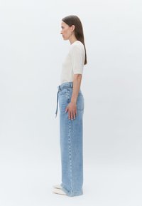 Haut blanc ajusté à manches courtes, jean bleu clair taille haute et chaussures beiges à bout pointu. Les jeans sont dotés d'une ceinture nouée et de poches.