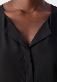 Camicetta nera con una texture morbida, caratterizzata da un colletto a contrasto e una chiusura con bottoni. Il tessuto appare leggero e liscio.