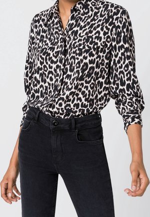 Vrouw draagt een zwart-wit shirt met luipaardprint en lange mouwen, ingestopt in een zwarte, hoogtaillespijkerbroek met zichtbare knopen en zakken.