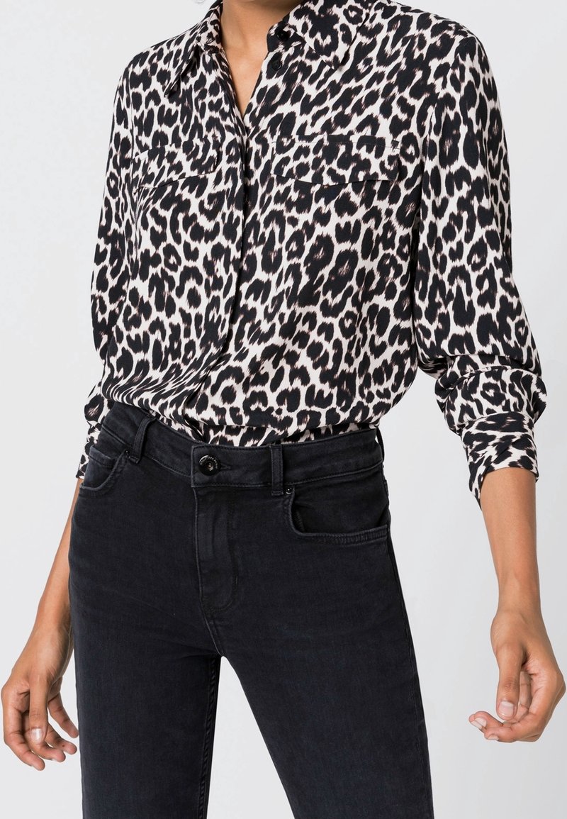 Femme portant une chemise à manches longues imprimé léopard noir et blanc, rentrée dans un jean noir taille haute avec des boutons et des poches visibles.
