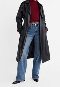 Trench coat nero in pelle con ampi rever, vita cintura, abbinato a jeans blu strappati a vita alta e stivali neri con punta a punta.