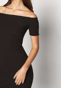 Svart avskuren klänning med korta ärmar, räfflad textur, figurnära silhuett och subtila fransar längs halsringningen och ärmarna.