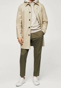 Trench coat beige chiaro indossato sopra un felpa grigia, abbinato a pantaloni verde oliva e sneaker bianche. Caratterizzato da chiusure a bottone e una vestibilità morbida.