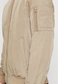 Beige Bomberjacke mit gerippten Bündchen und Saum, mit einer Reißverschlusstasche am Ärmel und zwei seitlichen Taschen. Glattes, leichtes Material.