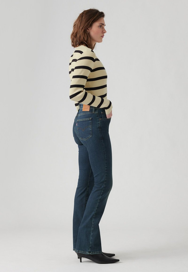 Suéter ribeteado a rayas en color crema y negro, combinado con unos jeans acampanados de un azul oscuro que tienen un parche con la marca en la parte trasera. Se completa con unos zapatos de tacón negro y puntera puntiaguda.