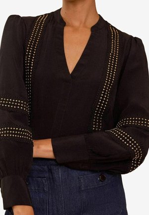 Blouse noire avec un col en V, accents texturés, clous dorés et détails en dentelle le long des manches, associée à un pantalon en denim foncé.