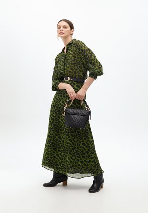 Maxikleid mit grünem Leopardenmuster und schwarzem Gürtel. Das Model hält eine kleine gewebte schwarze Handtasche mit Kettenakzenten. Schwarze Knöchelstiefel runden den Look ab.