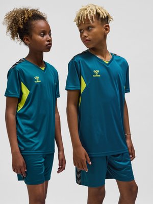 Zwei Kinder tragen passende türkis-gelbe Sporttrikots und Shorts und stehen nebeneinander vor einem einfarbigen Hintergrund.