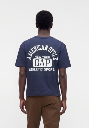 Muž otočený chrbtom, oblečený v tmavomodrom GAP tričku s textom "American Style New York GAP Athletic Sports" a v hnedej nohavici na jednoduchom pozadí.