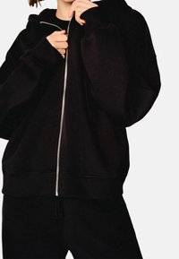 Czarna bluza z kapturem na zamek błyskawiczny, z dużym kapturem, ściągaczami na mankietach i u dołu, z metalowym zamkiem w kolorze srebrnym i luźnym krojem. Wykonana z miękkiej tkaniny.