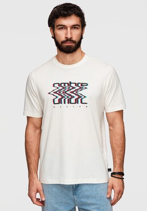 Homme barbu portant un t-shirt blanc avec un design de texte multicolore déformé et un jean bleu clair, debout devant un fond uni.