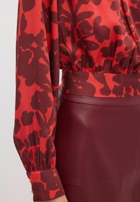 Rode blouse met bloemmotief en lange, opgeblazen mouwen, en een elastische tailleband. Gepaard met een donkere bordeauxkleurige high-waisted kunstlederen rok.