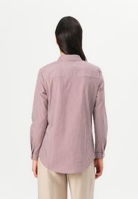 Femme aux longs cheveux foncés portant une chemise à manches longues à rayures verticales rouges et blanches et un pantalon beige, vue de dos sur fond blanc.
