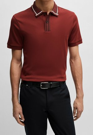 Mężczyzna w rdzawej koszulce polo z białym wykończeniem na kołnierzyku, czarnym paskiem i czarnych spodniach, stojący na jednolitym tle.