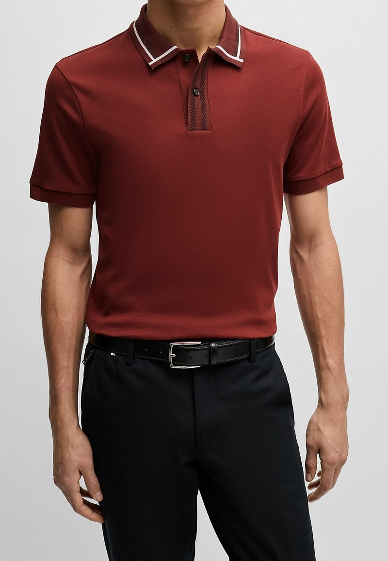 Homme portant un polo couleur rouille avec un col orné de liserés blancs, une ceinture noire et un pantalon noir, debout devant un fond uni.