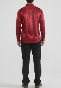 Chemise rouge en satin à boutons, à manches longues, avec empiècement au dos et poignets. Associée à un pantalon noir et des chaussures sombres. Texture lisse, finition brillante.