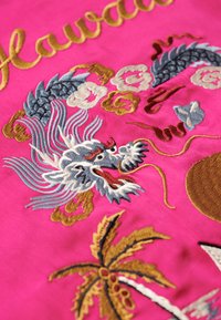 Tissu rose avec une broderie détaillée représentant un dragon, des palmiers et des motifs floraux en fils dorés, bleus et blancs. Texte "Hawaii" en doré.