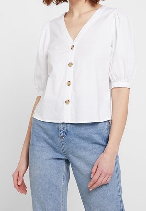 Kvinde iført en hvid V-hals bluse med puffede halværmer og fire brune knapper, kombineret med lyseblå, højtaljede jeans.