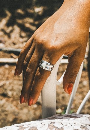 Hånd med rød neglelak bærer en sølv dobbeltbåndsring med en rund hvid sten og hviler på en hvid metalstol udendørs.