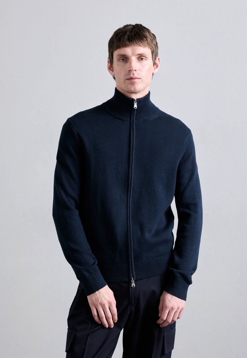 Mornarsko modra zip-up jopica z visokim ovratnikom, rebrastimi manšetami in robom. Narejena iz gladkega pletenega materiala, ima srebrno zadrgo.