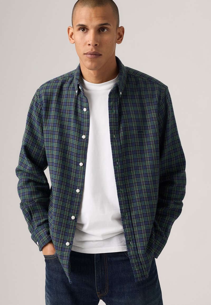 Man wearing unbuttoned dark green and blue plaid shirt over white t-shirt, with hands in pockets and neutral background.