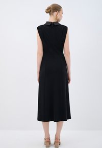 Robe noire avec des manches courtes structurées, un col et une coupe mi-longue. Texture lisse avec de subtiles plis sur les côtés et un zip au dos.